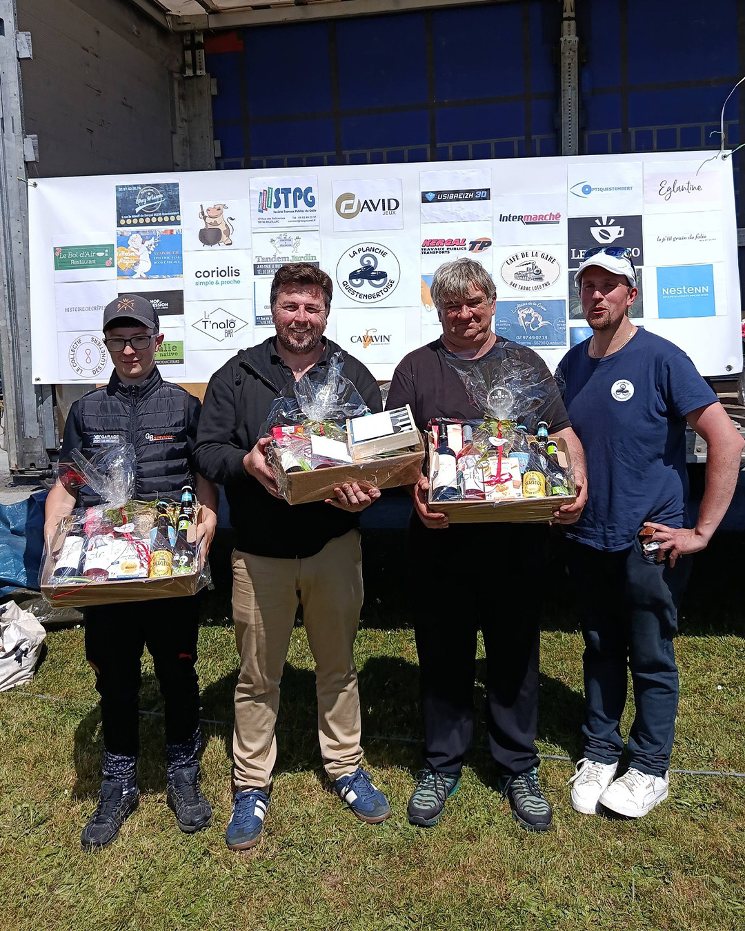 Four men stand on grass holding gift baskets wrapped in cellophane at an event podium with sponsor logos in the background, smiling for the photo.