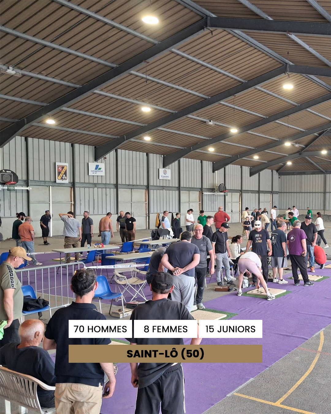 Indoor sports hall with many people standing and sitting around purple mats, watching a competition; banner reads Saint-Lô (50).