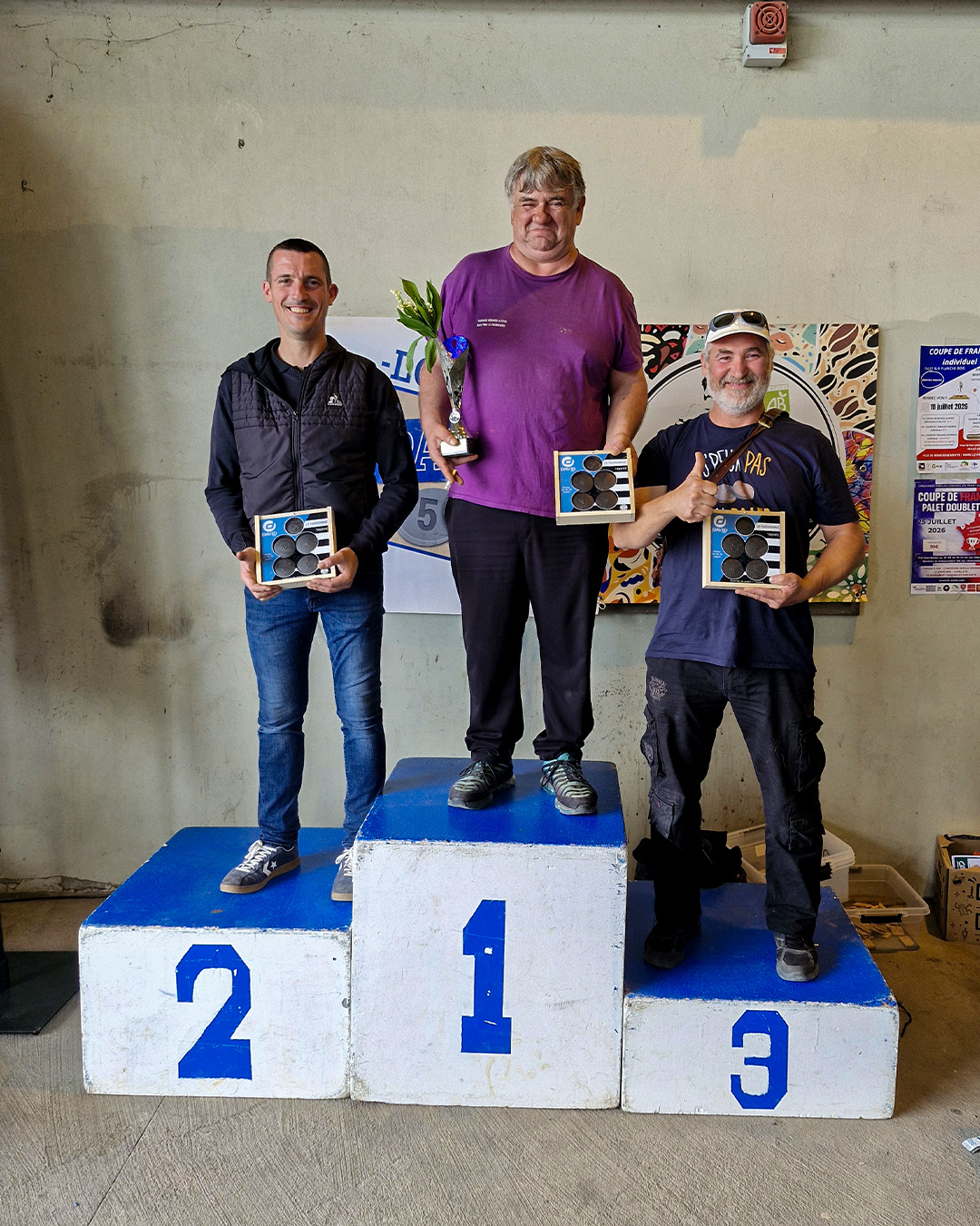 Three men stand on a winners' podium indoors; center is first place with a trophy, flanked by second and third place holding boxes.