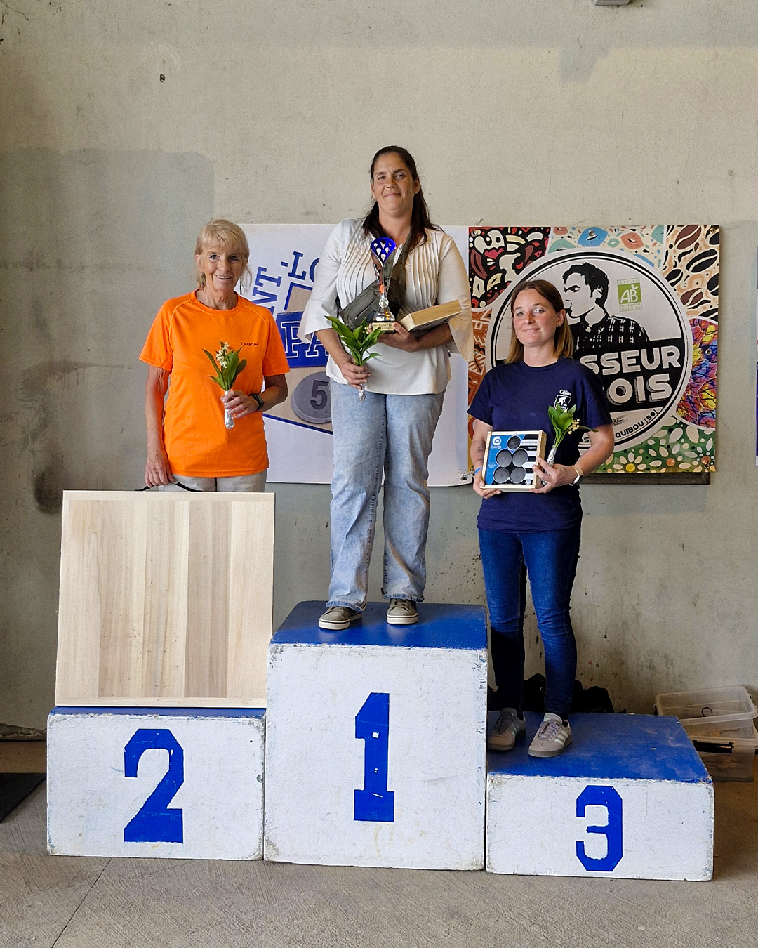 Three women stand on a winners podium indoors: first place in the center with a trophy and plant, second place to the left with a flower, third place to the right with a small prize and flower.