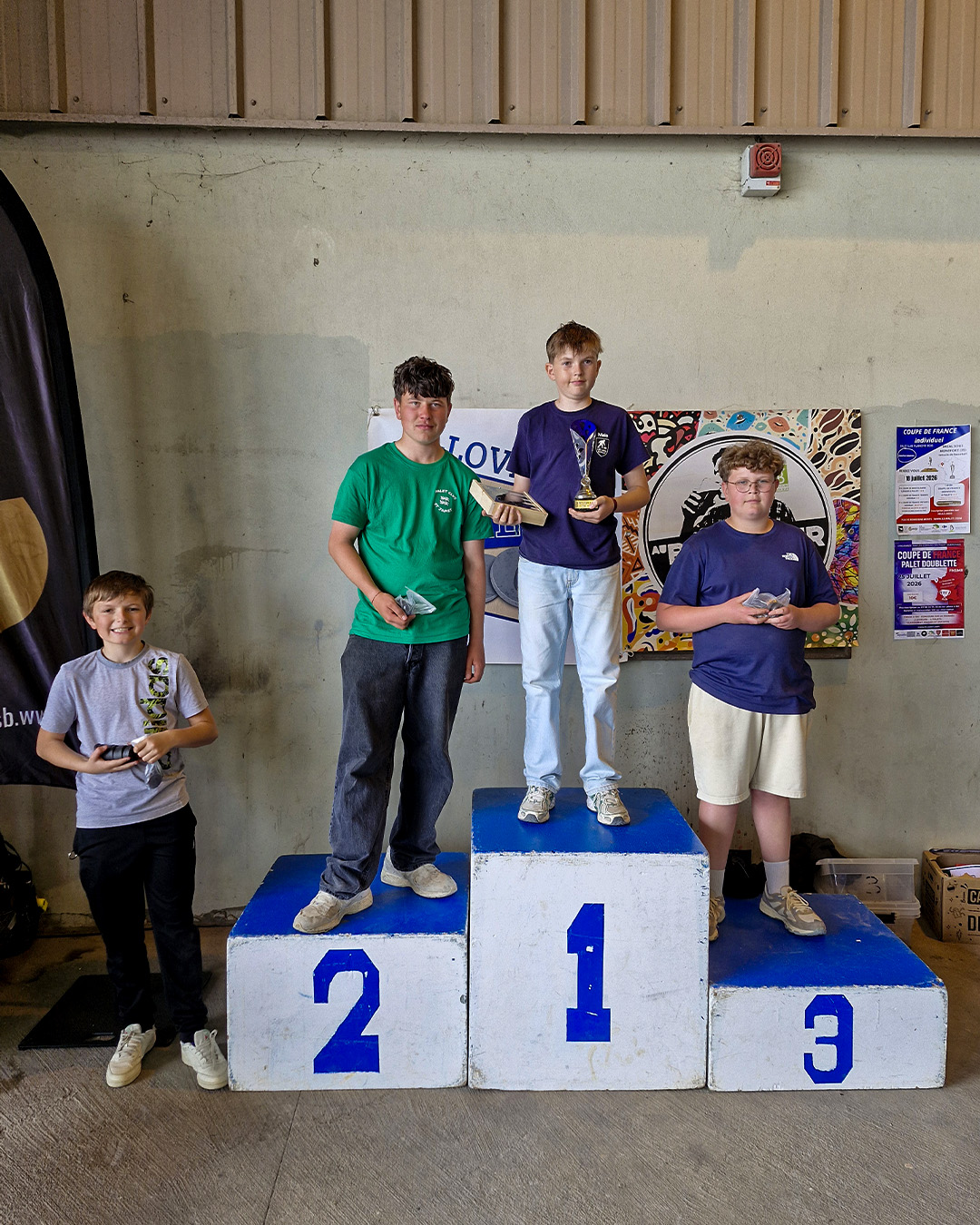 Three boys stand on a victory podium indoors, with the center winner in first place holding a trophy between second and third places on either side.