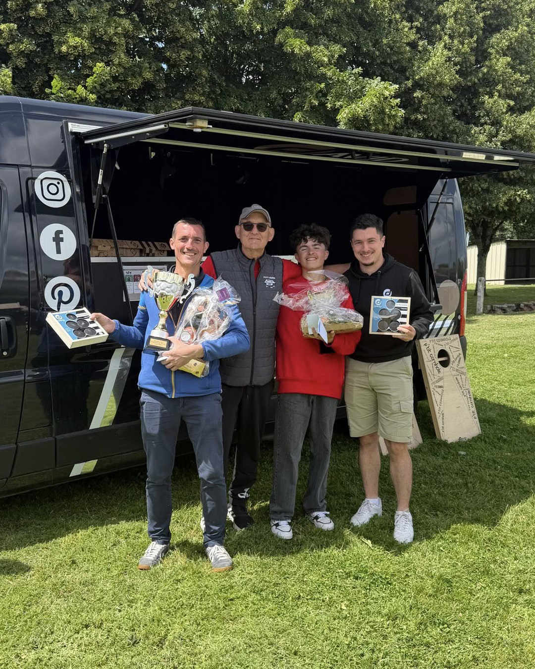 Four men pose with trophies and gift baskets in front of a dark van at an outdoor awards event on a grassy field.