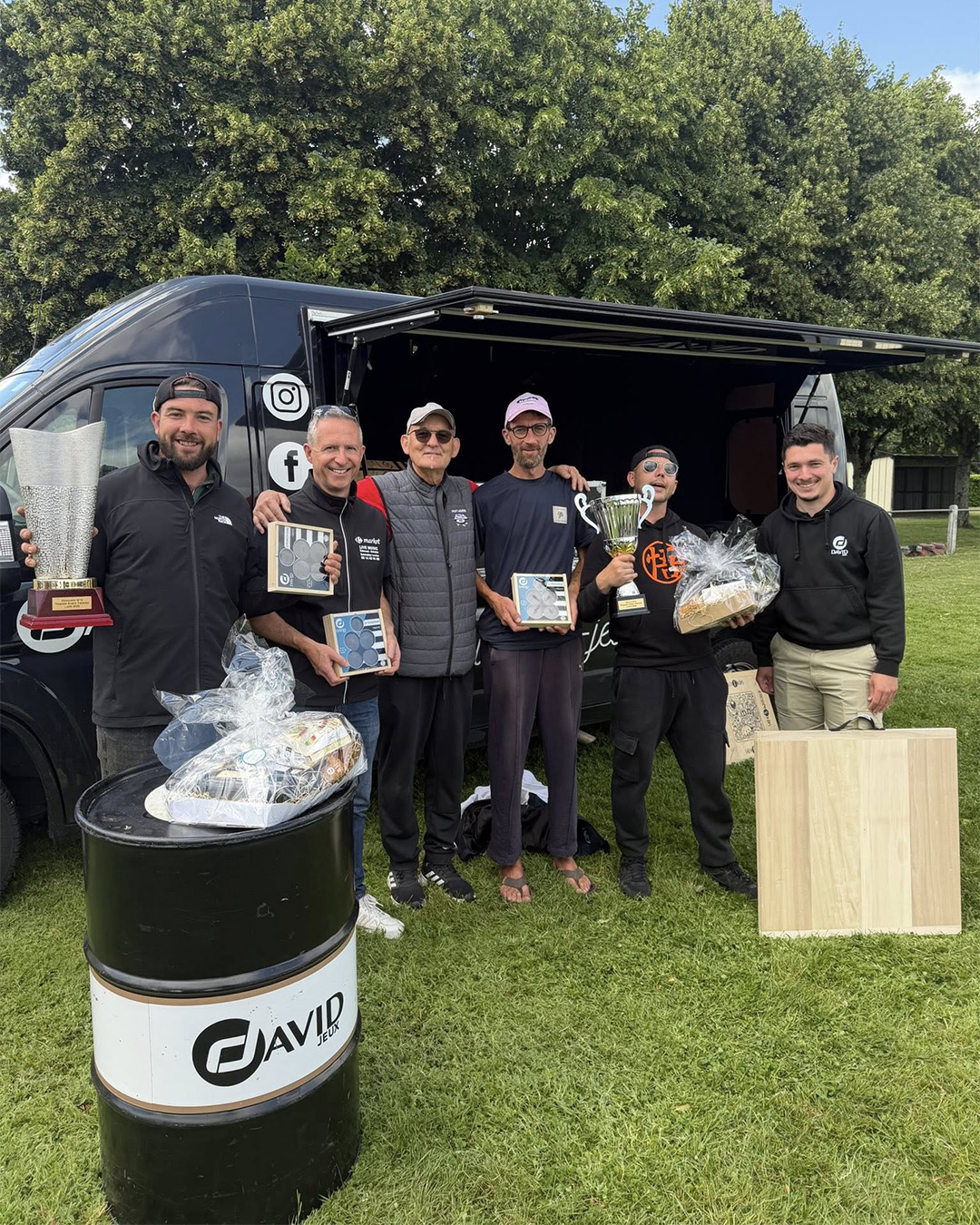 Group of men posing with trophies and gift baskets in front of a black van, outdoors at an awards event.
