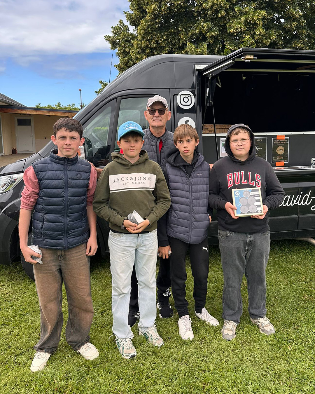 Five boys and an older man pose in front of a dark van on a grassy field; the boy on the far right holds a boxed item.