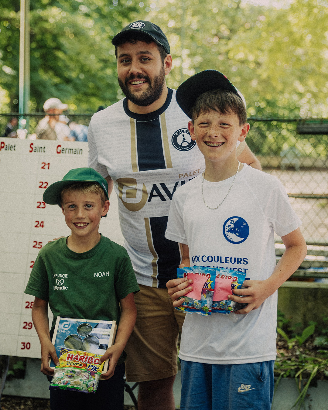Three smiling boys posing for a photo at an outdoor sports event, each holding prize candy bags and wearing caps and casual clothes, with a scoreboard in the background.