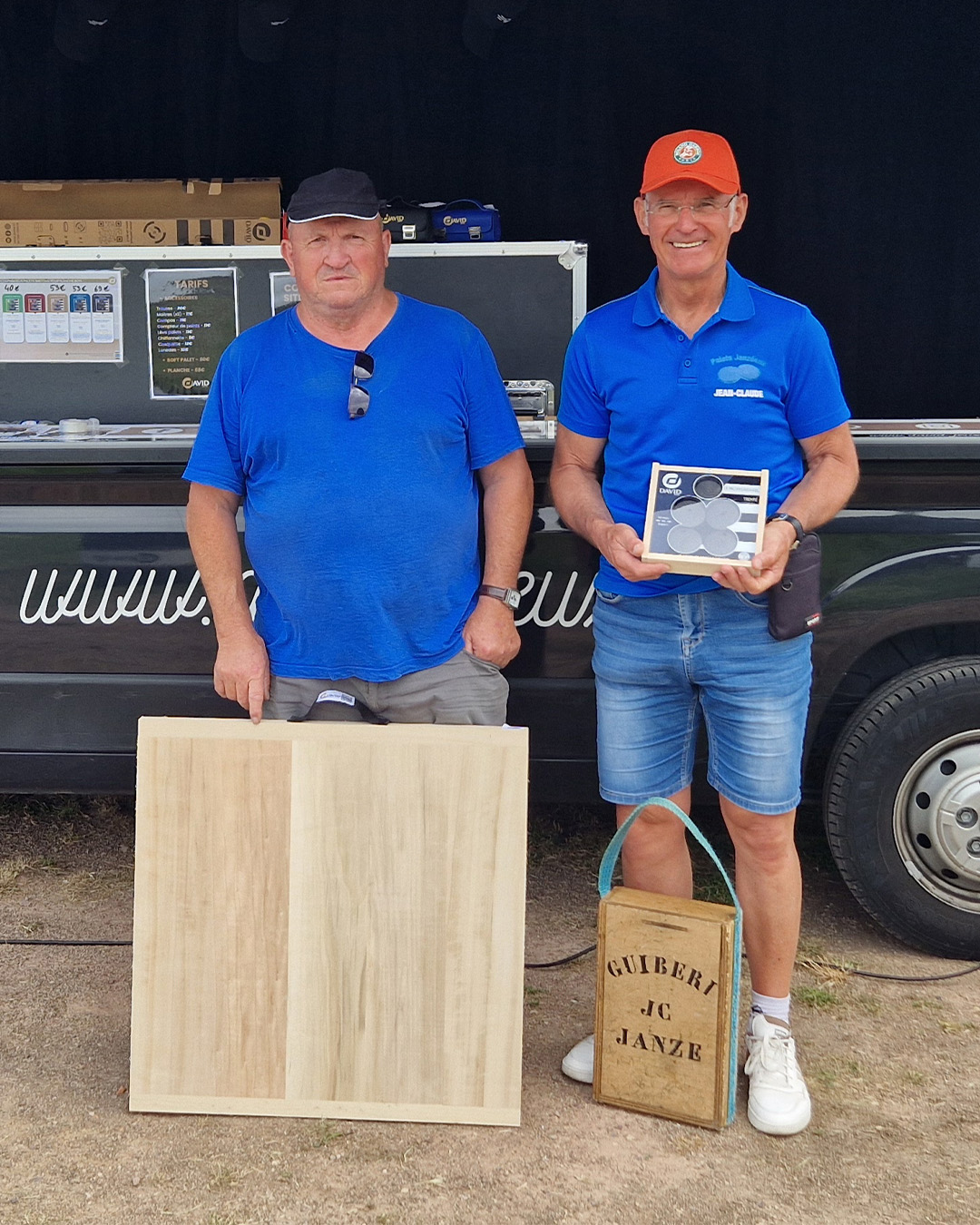Two men in blue shirts stand beside a service trailer; the man on the right holds a display box with circular components.